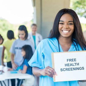 Confident African American female nurse holds 'Free Health Screenings' sign to promote health expo. Patients and doctors are in the background.